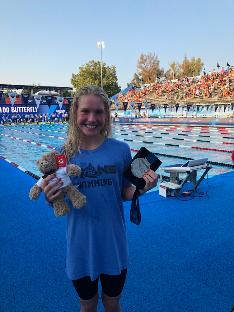 14-year-old Claire Curzan smiles in front of the pool, holding the medal and teddy bear she was awarded as prizes.