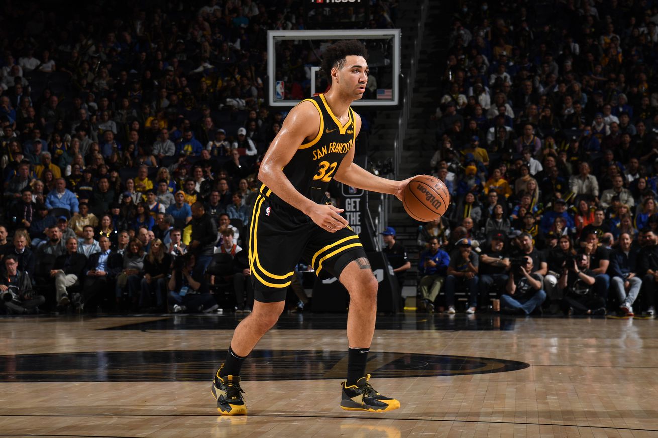 Trayce Jackson-Davis dribbles the ball to the left during the game against the Memphis Grizzlies at the Chase Center three-point line as the crowd cheers him on. The Warriors are wearing their black City Edition jerseys, and Jackson-Davis is wearing black-and-gold shoes which match his uniform.