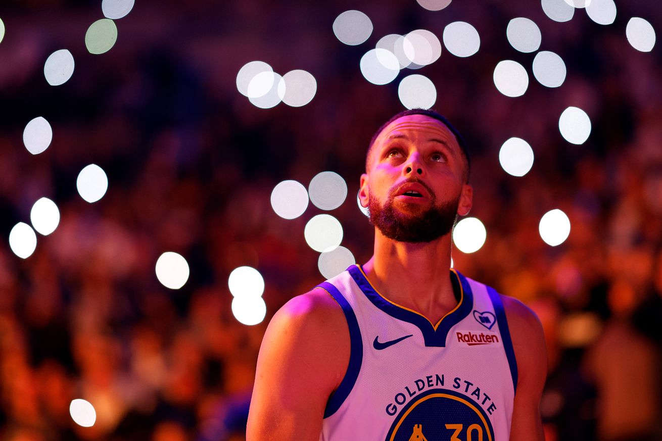 Stephen Curry looks on prior to the start of the game against at Target Center. He is surrounded by bokeh lights in a darkened arena. He is wearing a white uniform.