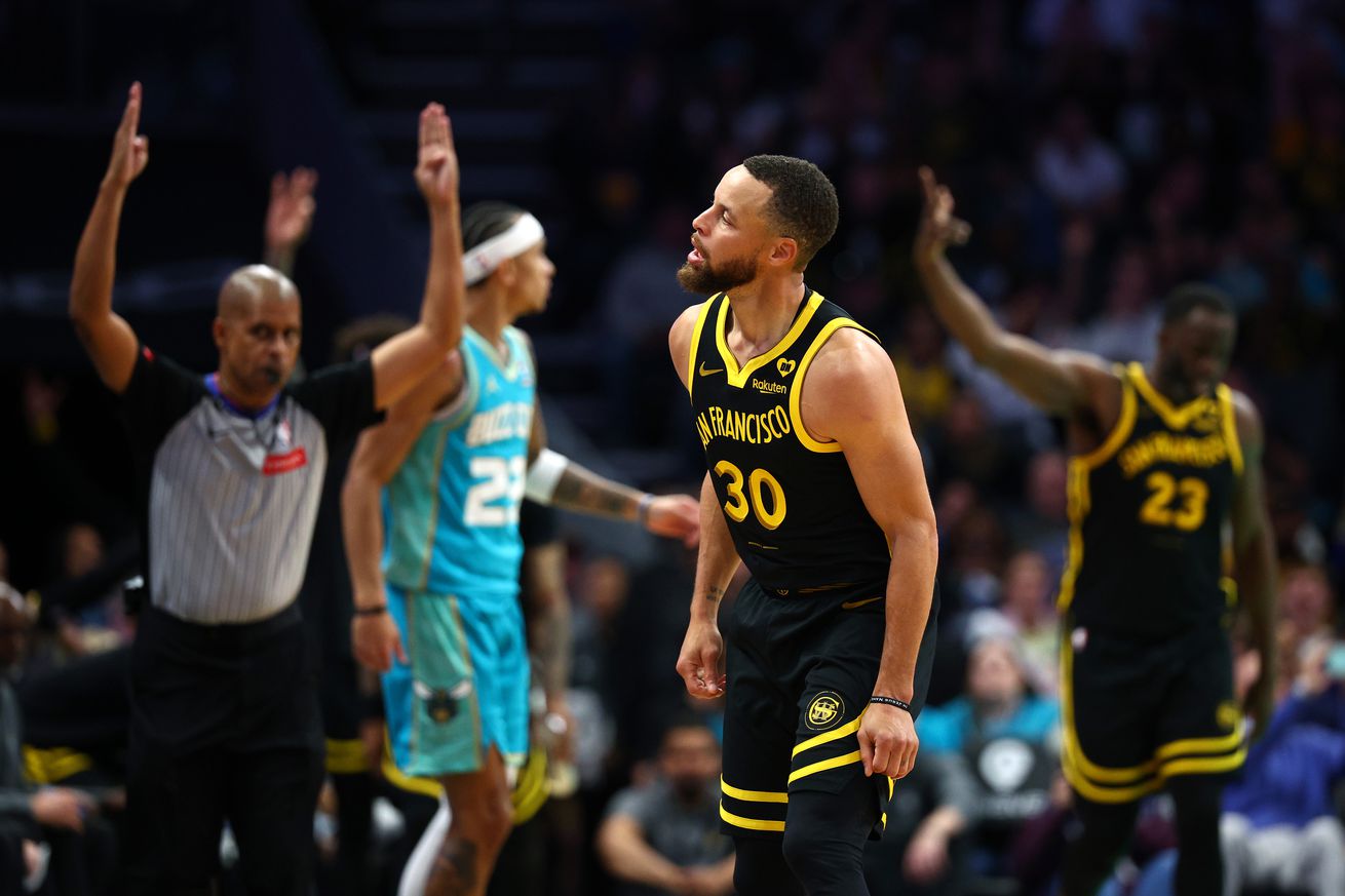 Steph Curry celebrating as he runs down the court, with a ref and Draymond Green behind him, both signaling a three. 