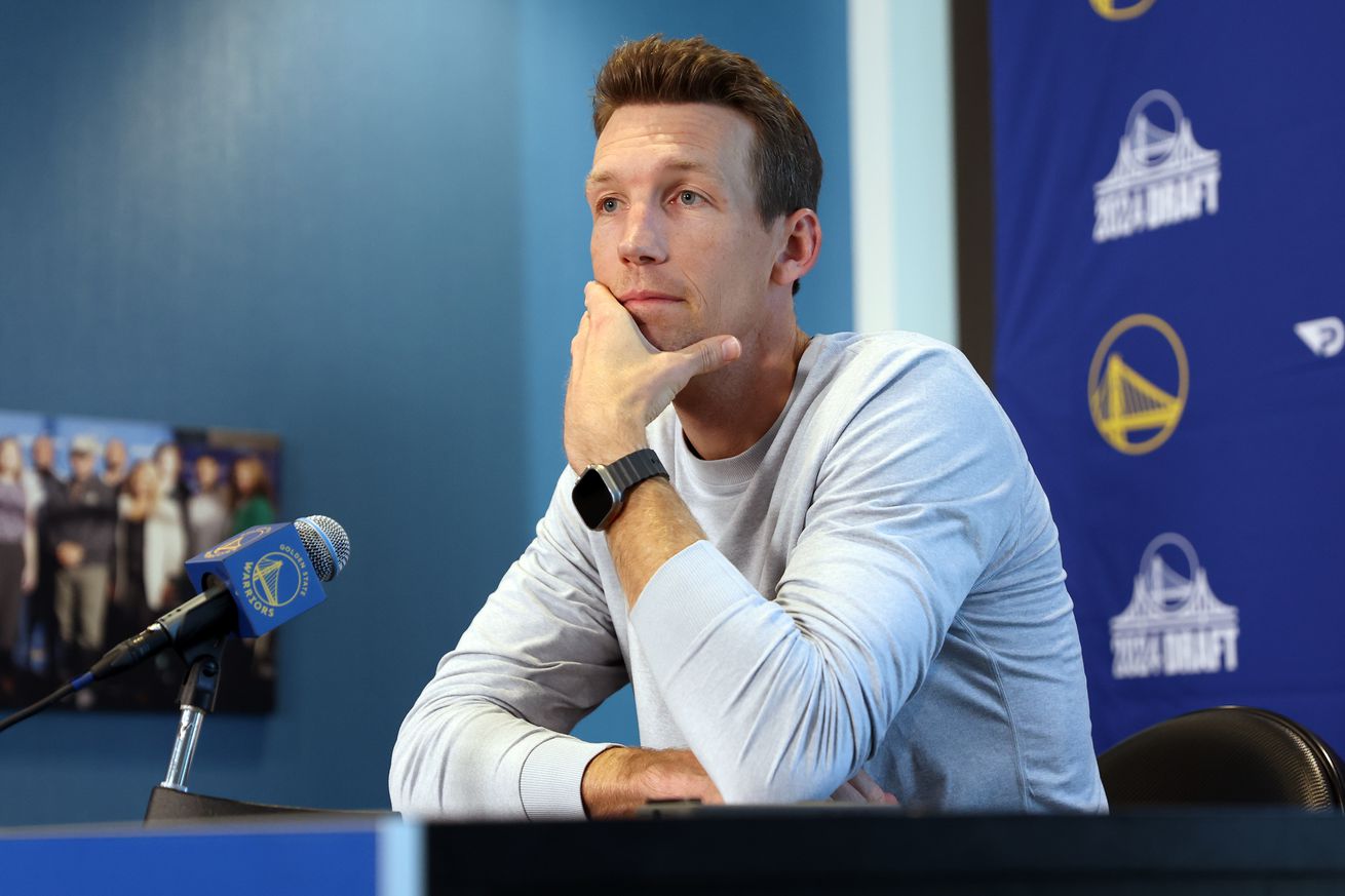 Mike Dunleavy Jr. sitting at a podium, with a Warriors background behind him, and his hand on his chin. 