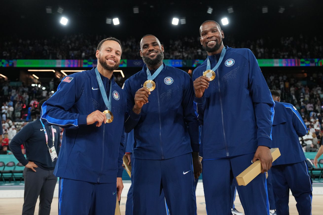 Stephen Curry #4, LeBron James #6, and Kevin Durant #7 of Team USA pose for a photo in the arena after winning the gold medal. They are all smiling and holding their gold medals to the camera.