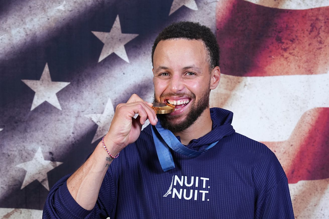 Stephen Curry of the USA Men’s National Team poses for portraits with the Gold Medal at the hotel on August 10, 2024 in Paris, France. He is smiling and biting the medal. He is standing in front of an American flag backdrop. He is wearing a dark blue ‘nuit nuit’ hoodie.