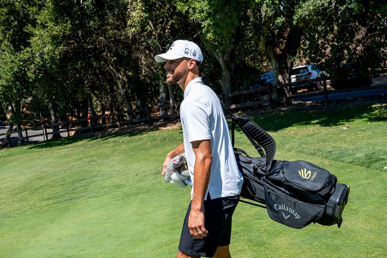 Steph Curry walking on a golf course with a bag of clubs. 