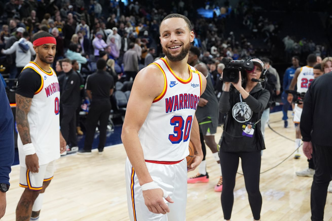 Steph Curry smiling after a game, with cameras and Gary Payton II behind him. 