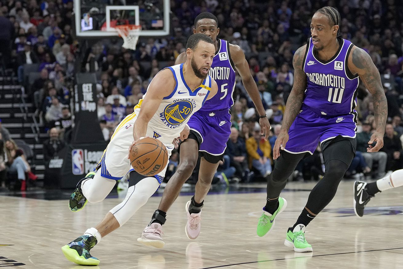 Steph Curry driving with De’Aaron Fox and DeMar DeRozan behind him. 