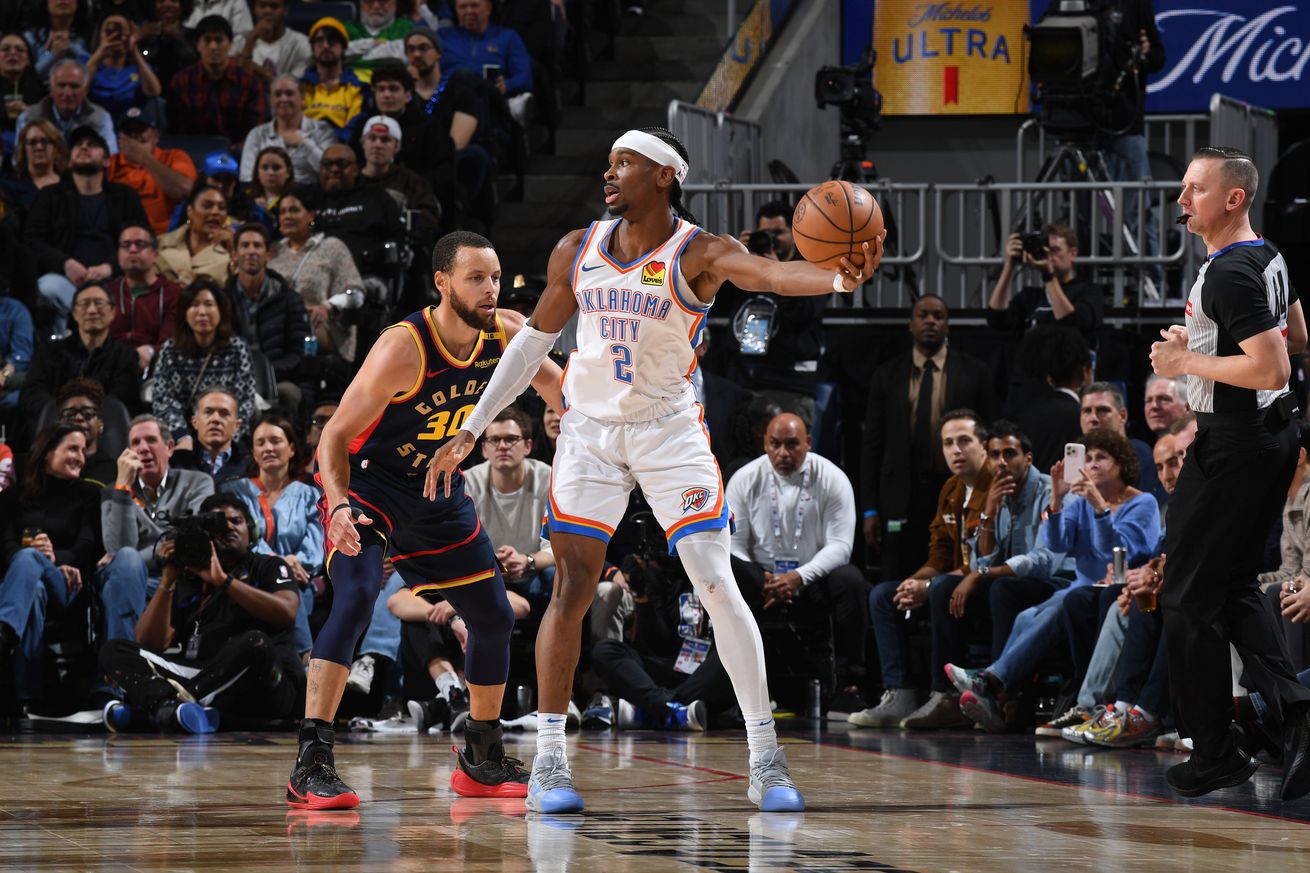 Shai Gilgeous-Alexander holding the ball while defended by Steph Curry. 