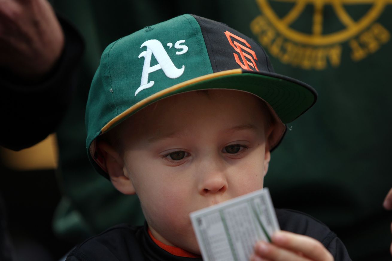 A young kid wearing a split Giants and A’s hat, sitting in a seat at a game, looking at a baseball card. 