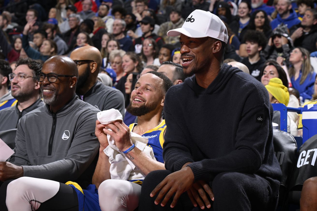 Jimmy Butler III, in street clothes, laughing on the bench with Steph Curry. 