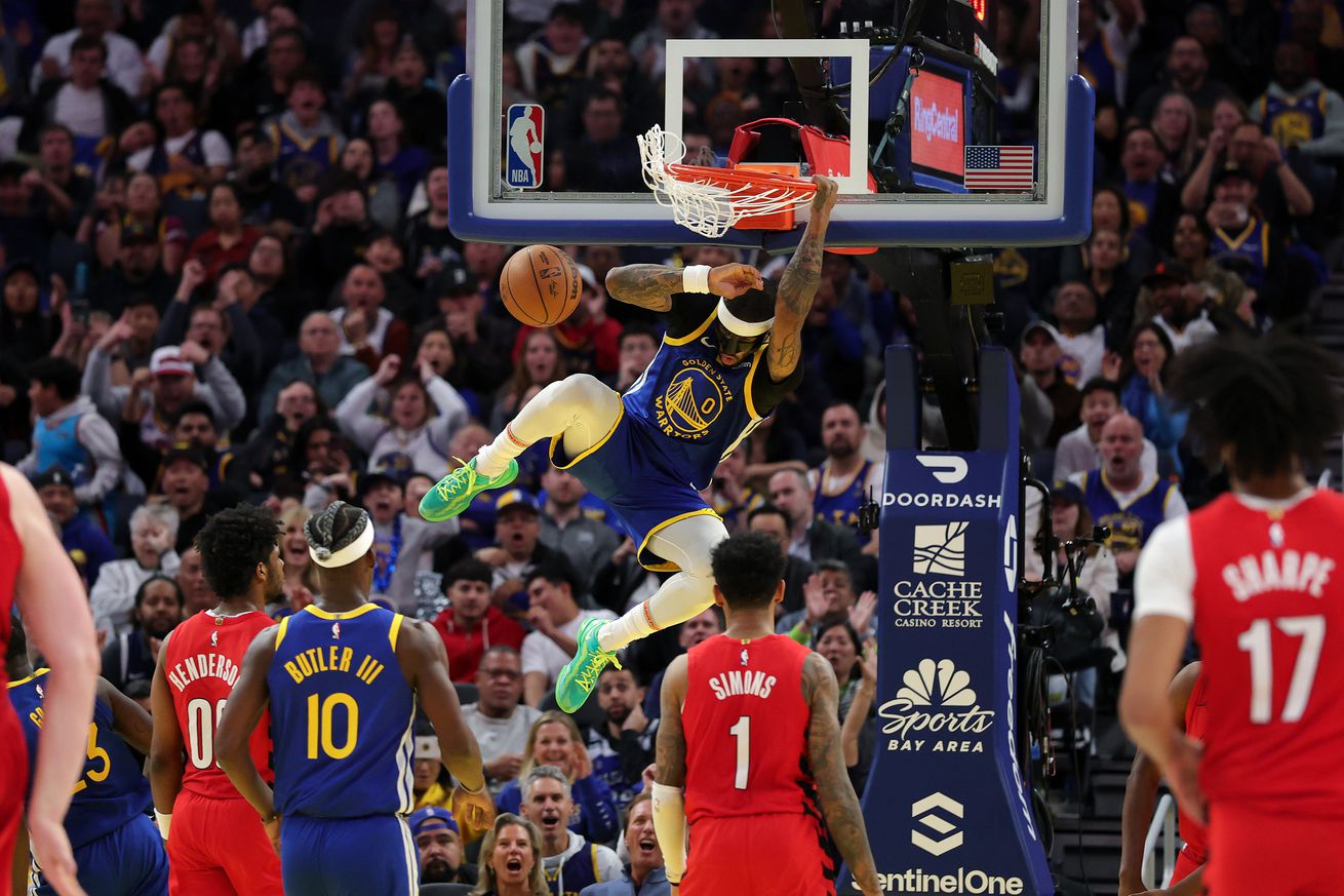 Gary Payton II hanging on the rim after a dunk, surrounded by Warriors and Blazers players. 